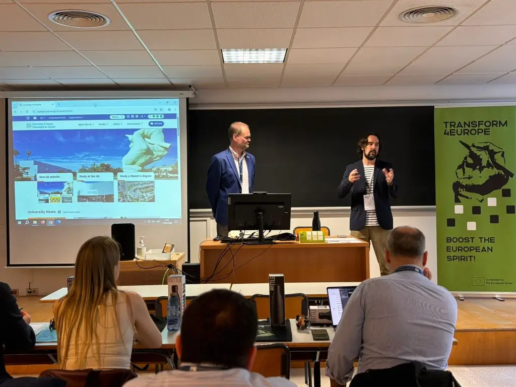 Two men present in a classroom with a projector screen displaying a website. Attendees sit at desks with laptops. A green “Transform4Europe” banner stands beside them. Classroom has white walls and a blackboard.