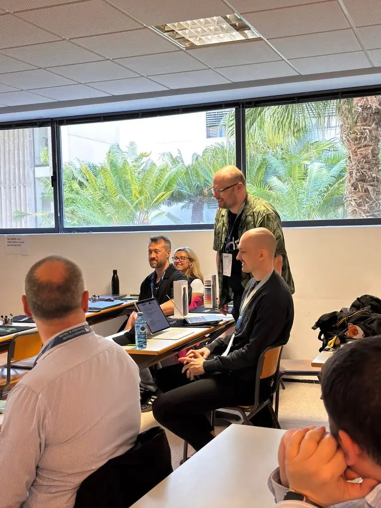 A group of people sits and stands in a classroom with large windows and palm trees outside. Some are using laptops, and they appear to be engaged in a discussion or presentation.