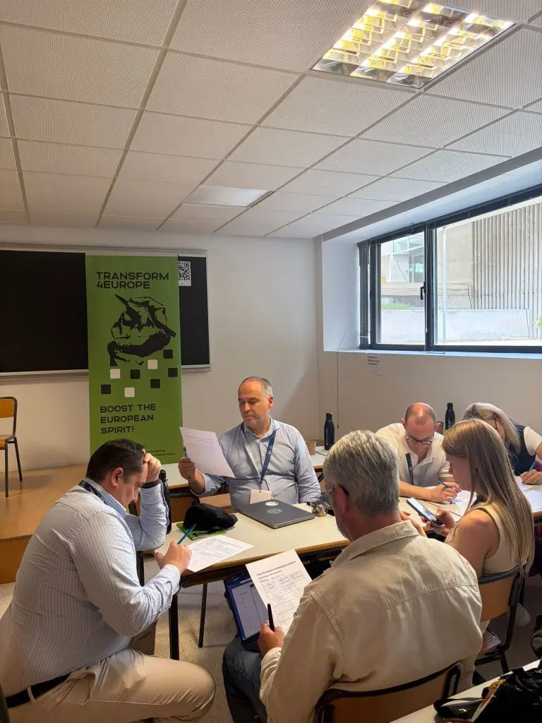 A group of people sit around a table in a classroom, engaged in discussion and reading documents. A green banner behind them reads “TRANSFORM EUROPE BOOST THE EUROPEAN SPIRIT!” Large windows let in daylight.
