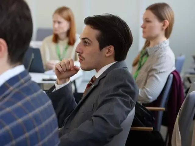 A young man in a suit sits attentively in a conference room, with other attendees visible in the background. They all appear focused on a presentation or discussion.