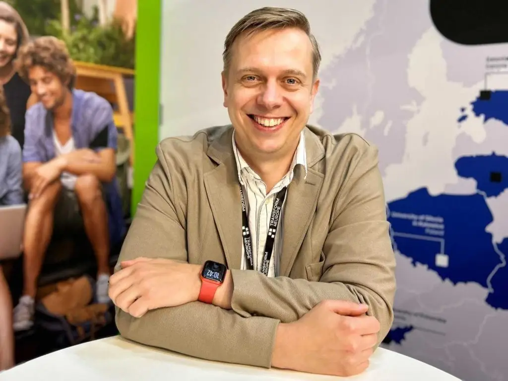 A smiling man with arms crossed sits at a table, wearing a beige jacket and a smartwatch. Behind him is a map of Europe with highlighted areas and a blurred image of seated people.