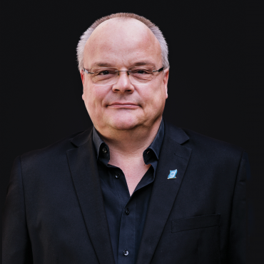 A middle-aged man with glasses and short gray hair is wearing a black suit jacket and black shirt, standing against a solid black background.