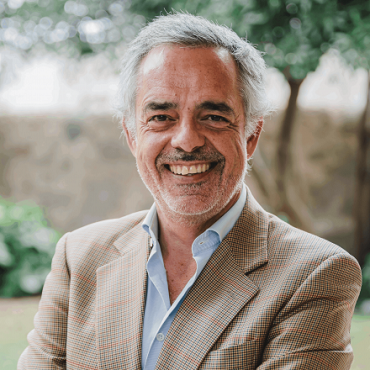 A middle-aged man with gray hair and a beard, wearing a checked beige blazer and blue shirt, smiles warmly while standing outdoors in a garden setting.