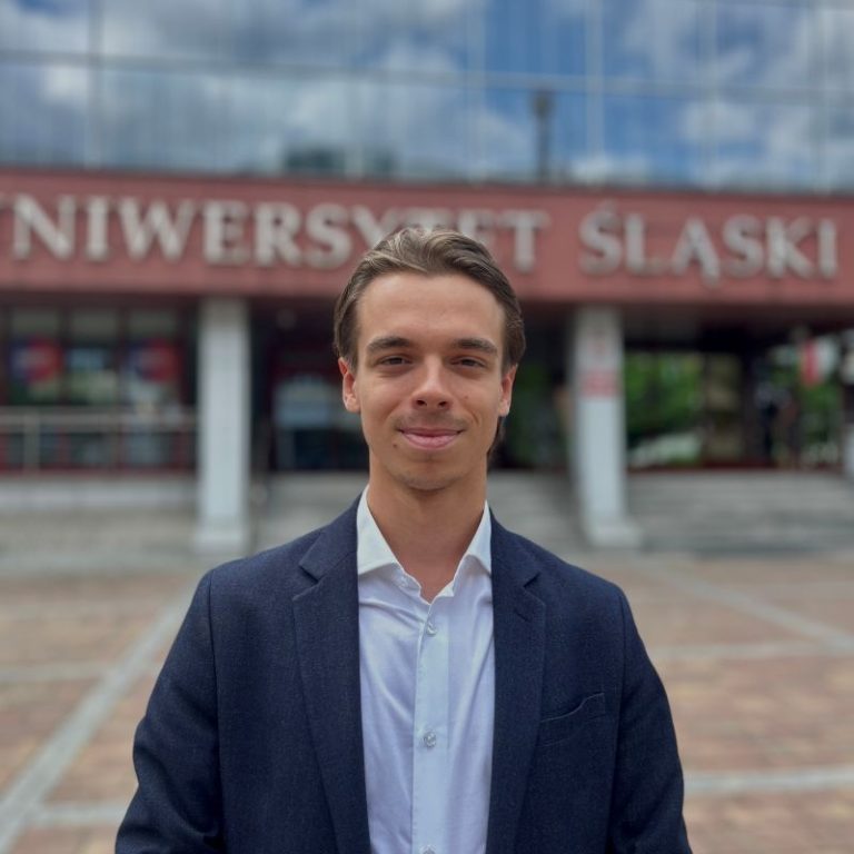 A young man in a navy blazer and white shirt stands smiling in front of a building with the sign UNIWERSYTET ŚLĄSKI, showcasing his pride as part of the Transform for Europe alliance. The background includes steps and large windows.