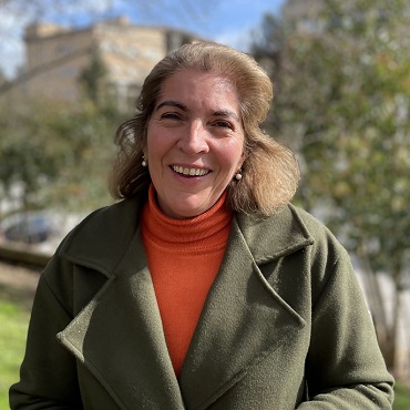 A woman with shoulder-length light brown hair, wearing an orange turtleneck and an olive green coat, smiles outdoors with greenery and buildings in the blurred background.