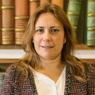 A woman with light brown hair, wearing a tweed jacket and white blouse, stands in front of a bookshelf filled with large, colorful books. She is looking at the camera and smiling slightly.