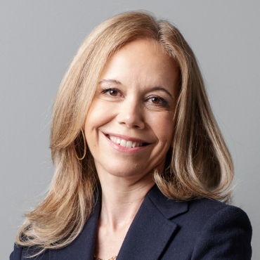 A woman with long blonde hair, wearing a dark blazer, smiles confidently at the camera against a plain gray background.