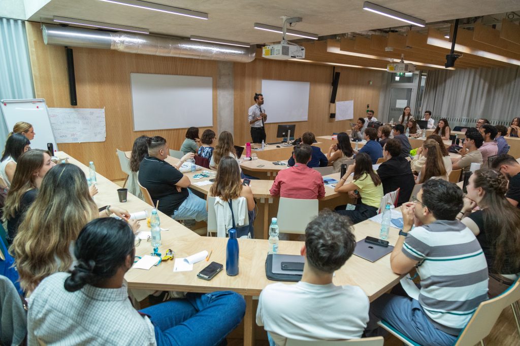 A group of people sit around tables in a modern classroom, listening to a person standing and speaking at the front. There are notebooks, water bottles, and laptops on the tables.