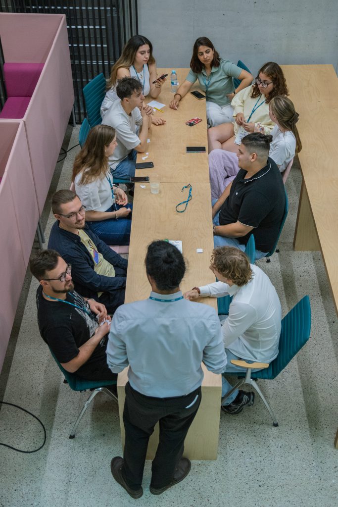 Twelve people gather around a long wooden table, engaged in discussion. Some are seated, while one person stands at the head of the table, addressing the group. Laptops, notebooks, and drinks are on the table.