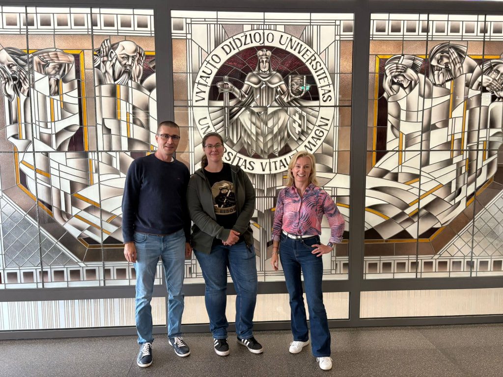Three people stand smiling in front of a stained glass wall featuring the seal of Vytautas Magnus University and abstract geometric designs.