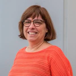 A woman with short brown hair and glasses smiles while wearing an orange textured top, standing in front of a plain light gray background.