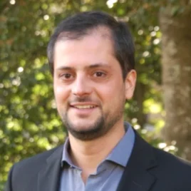 A man with short dark hair and a trimmed beard, wearing a dark blazer over a blue shirt, smiles outdoors with trees and greenery in the background.