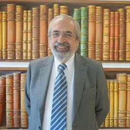 A smiling man with gray hair and a beard, wearing glasses, a gray suit, and a blue striped tie, stands in front of a bookshelf filled with large, colorful books.