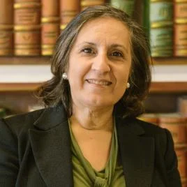A woman with shoulder-length gray hair, wearing a green blouse and dark blazer, smiles while sitting in front of bookshelves filled with large, colorful books.
