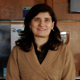 A woman with long dark hair, wearing a brown sweater and black shirt, stands indoors in front of a wooden wall with various plaques and papers displayed behind her.