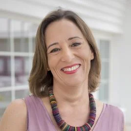 A woman with straight brown hair, wearing a sleeveless lavender top and a colorful beaded necklace, smiles at the camera. The background is bright with large windows.