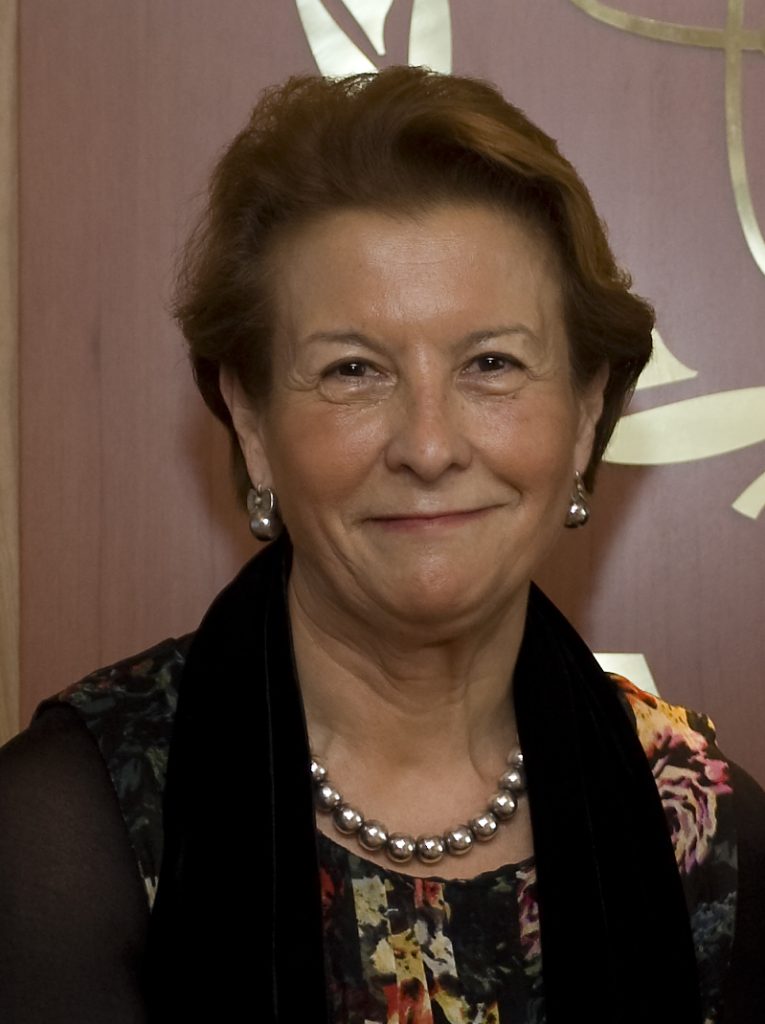 A woman with short brown hair wearing a floral dress, pearl necklace, and pearl earrings stands in front of a wooden background with gold detailing, smiling at the camera.
