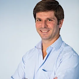 A man with short brown hair, wearing a light blue button-up shirt, stands smiling with arms crossed against a plain light blue background.