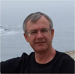 A middle-aged man with short gray hair and glasses sits outdoors near the ocean, wearing a black shirt. The background shows a cloudy sky and waves in the distance.