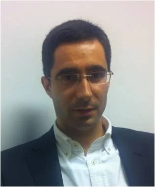 A man with short dark hair, glasses, and a serious expression is wearing a white button-up shirt and a dark blazer, seated against a plain light-colored wall.