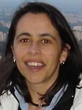 A woman with dark hair smiles at the camera, wearing a white and black jacket and earrings, with a blurred outdoor landscape in the background.