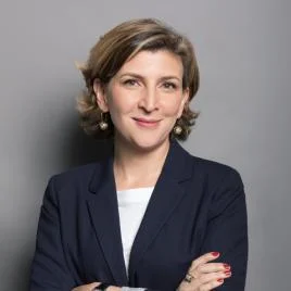 A woman with short, light brown hair wearing a navy blazer and white top stands with arms crossed, smiling confidently against a plain gray background.