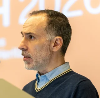 A man with short hair and a gray beard speaks at a podium with a microphone. He is wearing a collared shirt and sweater. The background is blurred with large white text on a peach-colored wall.