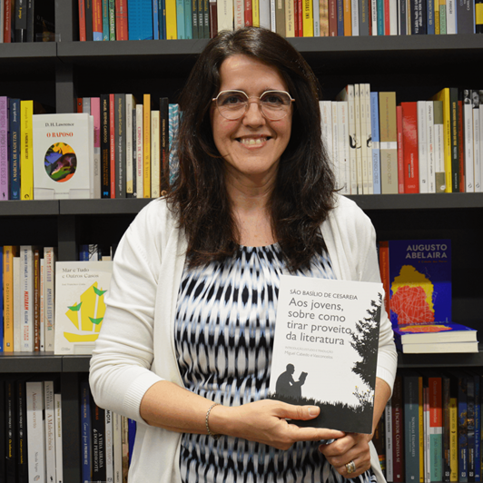 A woman with long dark hair and glasses smiles while holding a book in front of a bookshelf filled with colorful books.