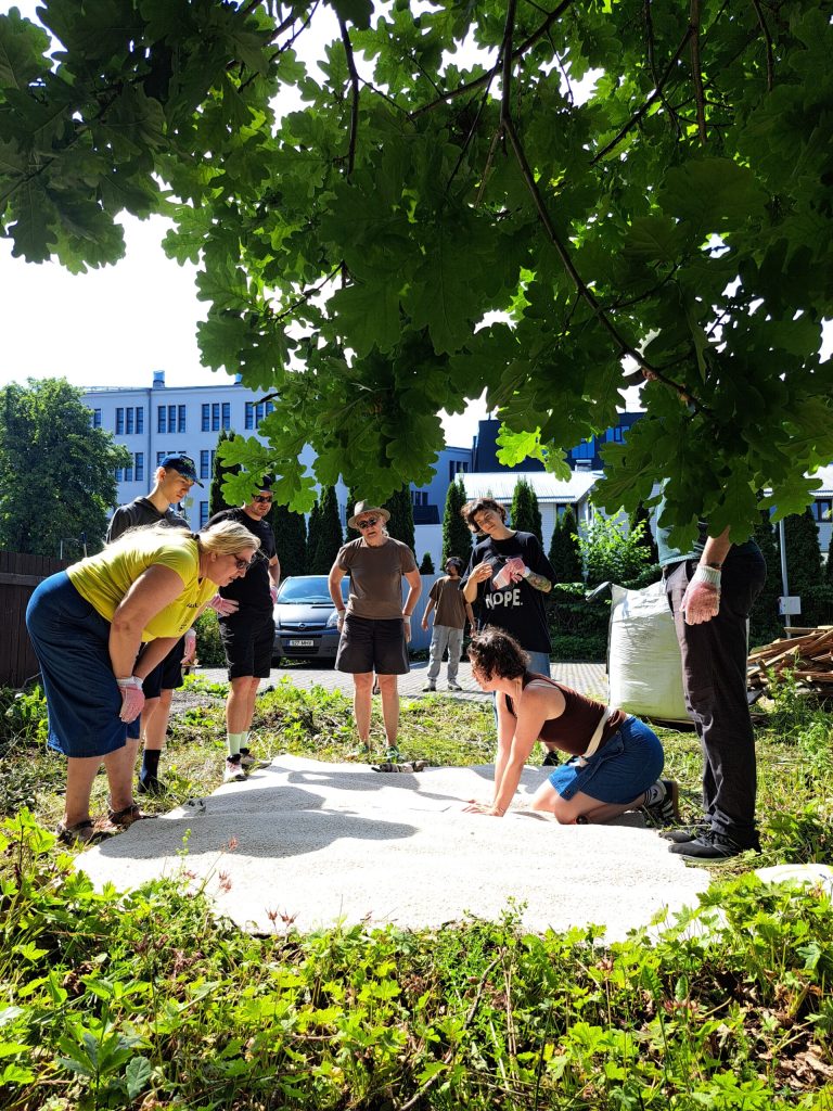 A group of people stand and watch as a woman kneels on the ground in a sunlit garden area, surrounded by greenery and overshadowed by large leaves, with buildings visible in the background.