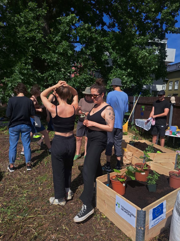A group of people gather outdoors around raised garden beds, some tending to plants in the sunshine. Two women in black clothing stand in the foreground, one adjusting her hair. Potted plants and gardening tools are visible.