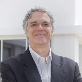 An older man with curly gray hair and glasses, wearing a dark suit and white shirt, stands smiling in front of a white building with windows.
