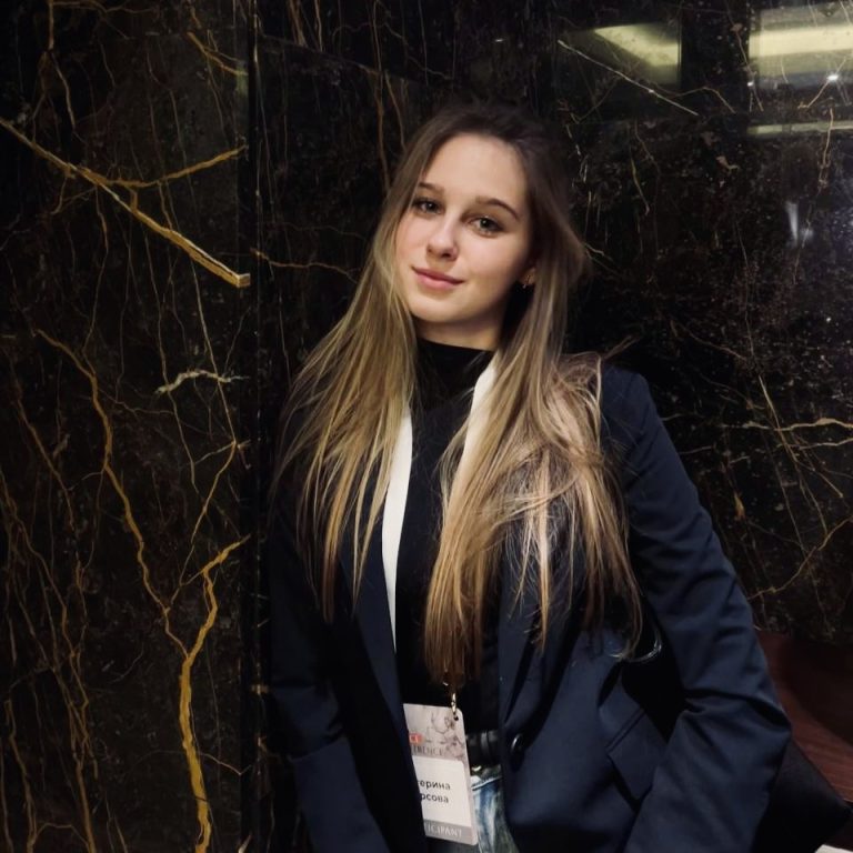 A young woman with long, straight blonde hair, wearing a dark blazer and a lanyard emblazoned with the "Transform for Europe" logo, stands in front of a dark marble wall with gold veining, looking at the camera and smiling softly.
