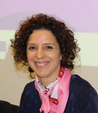 A woman with curly brown hair smiles at the camera. She is wearing a navy polka-dot top and a white scarf with pink and red details. The background is softly lit in pastel colors.
