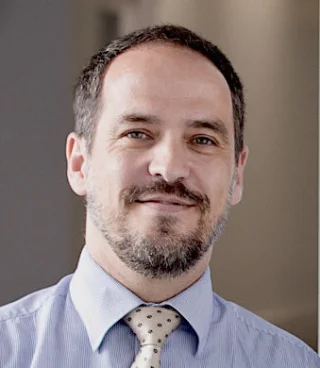 A man with short dark hair and a beard is smiling. He is wearing a light blue dress shirt and a cream-colored tie with small dark shapes. The background is softly blurred.