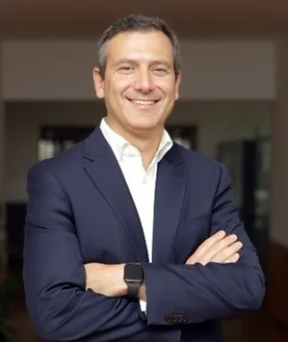 A man in a navy suit and white shirt stands indoors with arms crossed, smiling at the camera. He is wearing a smartwatch and appears confident and approachable.