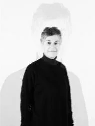 A grayscale image of a person with short hair wearing a dark top, standing in front of a light background. There is a faint, blurry duplicate silhouette behind them, creating a ghostly effect.