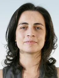 A woman with shoulder-length dark hair, light skin, and wearing a sleeveless dark top, looks at the camera with a neutral expression against a light background.