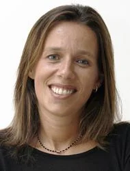 A woman with straight brown hair, wearing a black top and a beaded necklace, smiles at the camera against a plain white background.