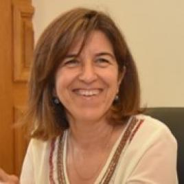A woman with shoulder-length brown hair, wearing a cream-colored blouse with embroidered details, is smiling while seated indoors near a wooden panel.