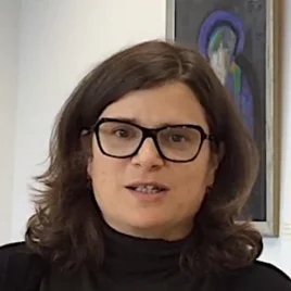 A person with shoulder-length brown hair and glasses wearing a black top stands indoors. A framed painting is visible in the background.
