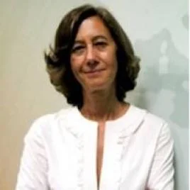 A woman with shoulder-length brown hair, wearing a white top, stands in front of a light-colored wall, smiling gently at the camera.