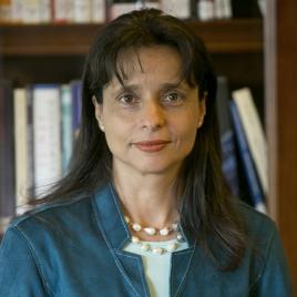 Woman with long dark hair, wearing a teal jacket and a pearl necklace, standing in front of a bookshelf filled with books.