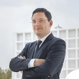 A man in a dark suit and tie stands with arms crossed, smiling slightly. He has short dark hair, glasses, and is outdoors with a modern white building in the background.