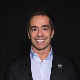 A man with short dark hair smiles, wearing a light blue shirt and a dark blazer against a plain black background.