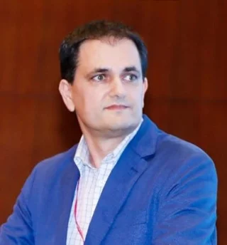 A man in a blue blazer and checked shirt looks to his side against a plain brown background.