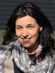 A person with dark hair wearing a patterned scarf smiles outdoors in sunlight, with a blurred background featuring horizontal lines.