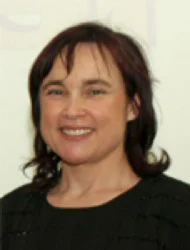 A woman with dark hair, wearing a black top, smiles at the camera in front of a light-colored background.
