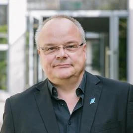 A middle-aged man with glasses and short hair, wearing a black suit and shirt, stands outside in front of a modern glass building, looking at the camera.