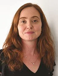 A woman with long, wavy red hair and fair skin wears a black top and a delicate necklace, standing against a plain white background, looking at the camera with a subtle smile.
