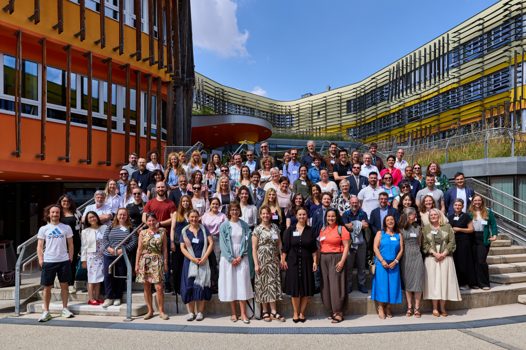 A large group of people pose together for a photo on outdoor steps in front of a modern, colorful building with glass and orange, yellow, and green panels under a blue sky.
